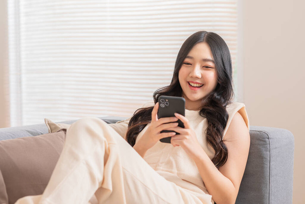 Happy asian young woman smile using smartphone surfing internet and enjoy with social media or online shopping at home. Modern wellness life of cute female looking at mobile phone fun and happiness - Фото, зображення