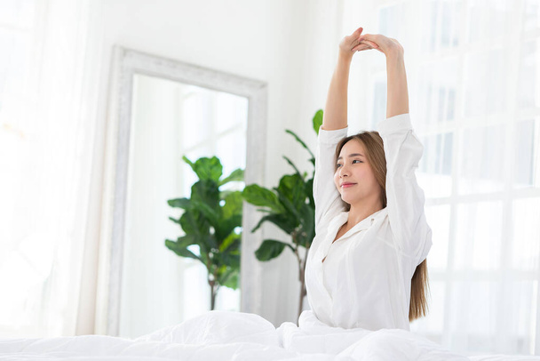 Hermosa mujer joven asiática estirando su brazo después de despertar en la cama y mirando las ventanas exteriores en el acogedor dormitorio sintiéndose tan fresca y relajarse por la mañana.Concepto de cuidado de la salud  - Foto, Imagen