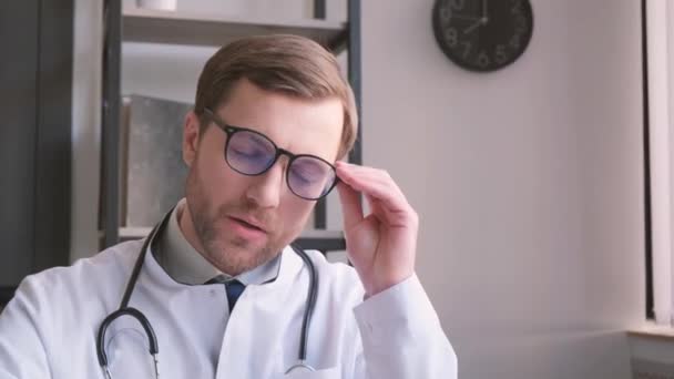 Κουρασμένος λυπημένος γενειοφόρος γιατρός σε λευκό παλτό απογειώνεται γυαλιά, κάθεται στο τραπέζι στο εσωτερικό του γραφείου. Προβλήματα στην εργασία, κόπωση στην κλινική. - Πλάνα, βίντεο