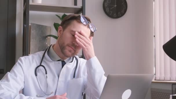 Deprimido joven médico con dolor de cabeza sentado en su oficina. Médico con exceso de trabajo en su consultorio.  - Metraje, vídeo