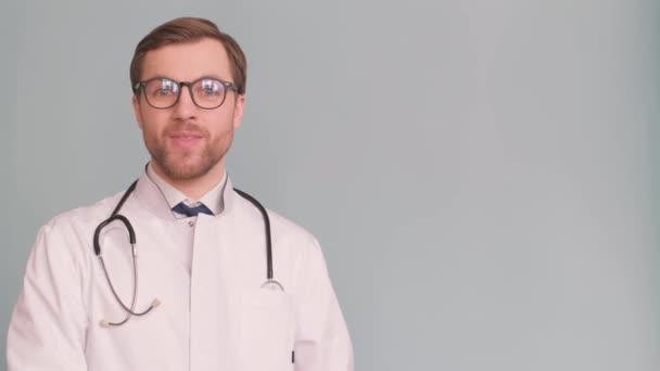 Χαμογελώντας νεαρός άνδρας γιατρός δείχνει και παρουσιάζει κάτι με το χέρι του, απομονωμένο σε γκρι φόντο. Υψηλής ποιότητας βίντεο 4k - Πλάνα, βίντεο