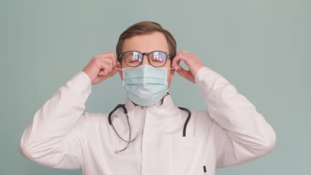 Retrato de un médico quitándose una máscara protectora, aislado sobre un fondo gris, está mirando a la cámara. Vídeo vertical - Metraje, vídeo