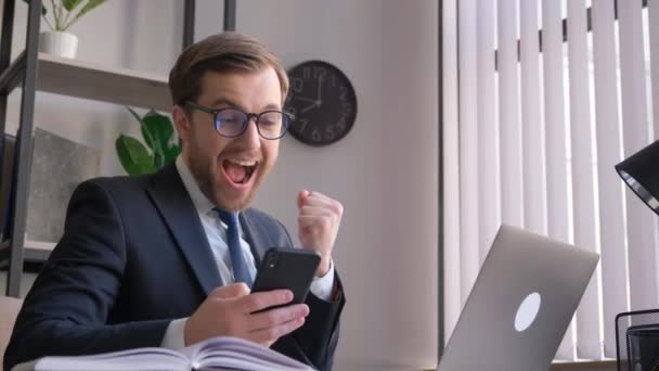 Grappige zakenman met smartphone in handen op kantoor die zich gelukkig en energiek voelt. Gekke kantoormedewerker in een staat van euforie - Video