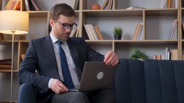 Caucasian young businessman working on laptop while sitting on sofa at home. The work of a freelancer. Free work schedule at home - Footage, Video