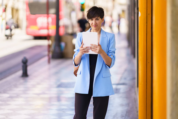 Koncentrált női vállalkozó formális ruhákban, táblagépen olvasva, miközben a városi utcán áll, és a képernyőt nézi homályos háttér az emberek és a busz nappal - Fotó, kép