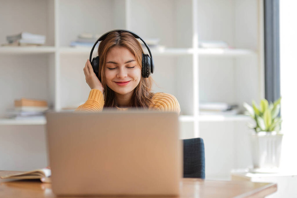ヘッドフォンをしているアジアの女性は、仕事中に音楽を聴きながらノートパソコンで楽しく音楽を聴いています。 - 写真・画像