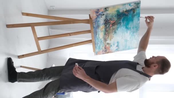Függőleges videó egy tehetséges művészről, aki ecsetet használ, hogy létrehozzon egy modern mesterművet egy olajfestményről egy fehér vásznon. A vászon egy állványon van a műhelyben. Függőleges videó - Felvétel, videó