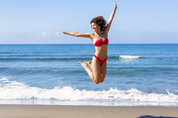 Возбужденная молодая женщина в розовом бикини вытягивает руки и прыгает в воздухе со сложенными ногами, улыбаясь и глядя в сторону при дневном свете на волнистое море и голубое небо - Фото, изображение