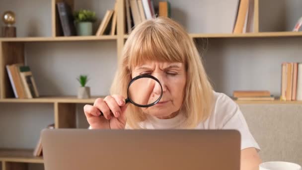 Egy idős nő egy nagyítón keresztül nézi a laptop képernyőjét. Egy gyönyörű szőke nagymama dolgozik egy laptopon a nappaliban. Függőleges videó - Felvétel, videó