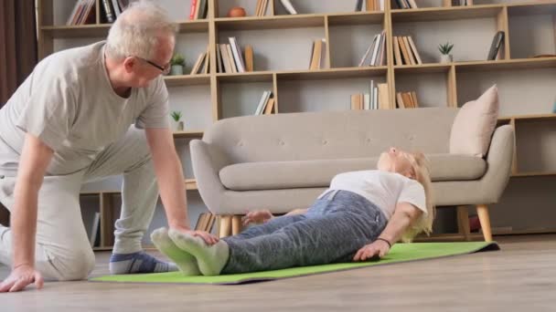 Happy elderly couple exercising at home. A successful grandmother pumps her abs with the help of her husband. Healthy lifestyle concept - Footage, Video