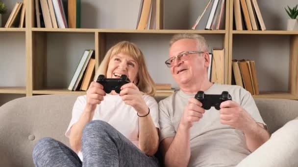 Pareja de ancianos muy felices jugando videojuegos mientras están sentados en la sala de estar. Entretenimiento para niños para personas mayores. Concepto de felicidad - Metraje, vídeo
