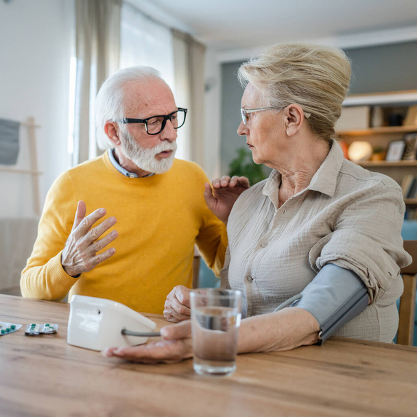 Vanhempi pari nainen tarkistaa mitata verenpainetta, kun hänen miehensä vanha valkoihoinen mies istuu pöydän vieressä kotona oikeat ihmiset perhe vanhukset eläkkeelle ja terveys käsite - Valokuva, kuva