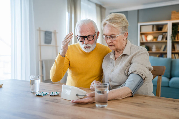 Oudere echtpaar controleren meten bloeddruk, terwijl haar man oude Kaukasische man naast de tafel zitten thuis echte mensen familie oude mensen pensioen en gezondheid concept - Foto, afbeelding