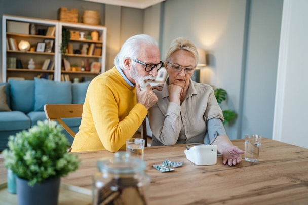 Starší pár žena kontrola měření krevního tlaku, zatímco její manžel starý běloch muž sedět vedle u stolu doma skuteční lidé rodina staří lidé odchod do důchodu a zdraví koncept - Fotografie, Obrázek