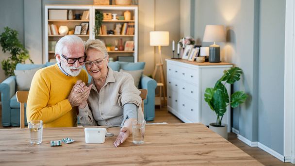 Senior couple woman check measure blood pressure while her husband old caucasian man sit beside at the table at home real people family old people retirement and health concept - Photo, image