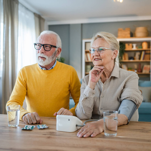 Senioren Paar Frau messen Blutdruck, während ihr Mann alten kaukasischen Mann sitzen neben am Tisch zu Hause echte Menschen Familie Senioren Ruhestand und Gesundheitskonzept - Foto, Bild