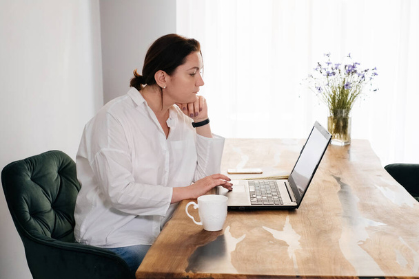 Middle age woman freelancer is working at home. Pretty plus size female in 40s working with laptop and phone from her kitchen in the morning with mug of hot drink. Real people lifestyle - Photo, Image