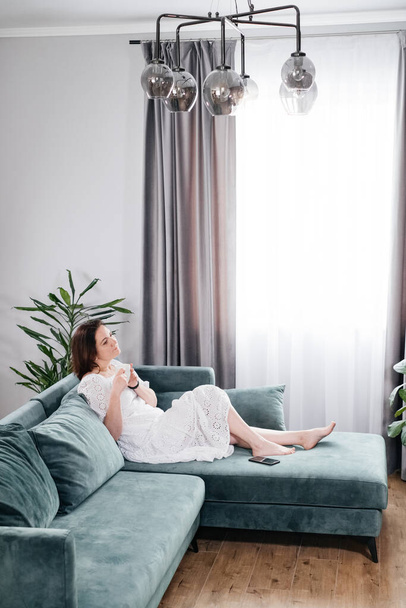 Woman drinking mug of coffee in cozy home atmosphere in the morning. Real middle age plus size female in white dress relaxing on the sofa, modern house. Sunny day with cup of tea, laying on couch - Fotoğraf, Görsel