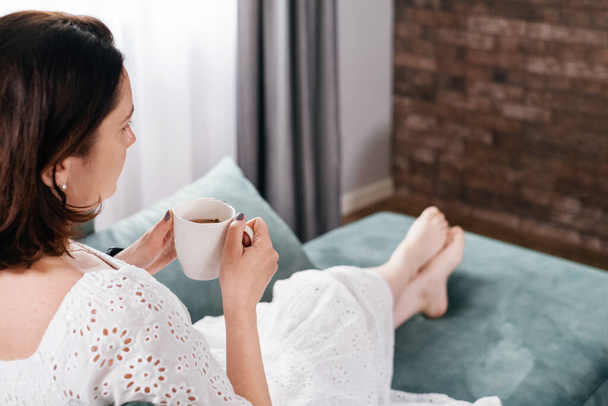 Woman drinking mug of coffee in cozy home atmosphere in the morning. Real middle age plus size female in white dress relaxing on the sofa, modern house. Sunny day with cup of tea, laying on couch - Photo, image