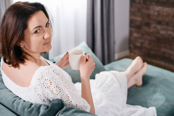 Woman drinking mug of coffee in cozy home atmosphere in the morning. Real middle age plus size female in white dress relaxing on the sofa, modern house. Sunny day with cup of tea, laying on couch - Foto, afbeelding