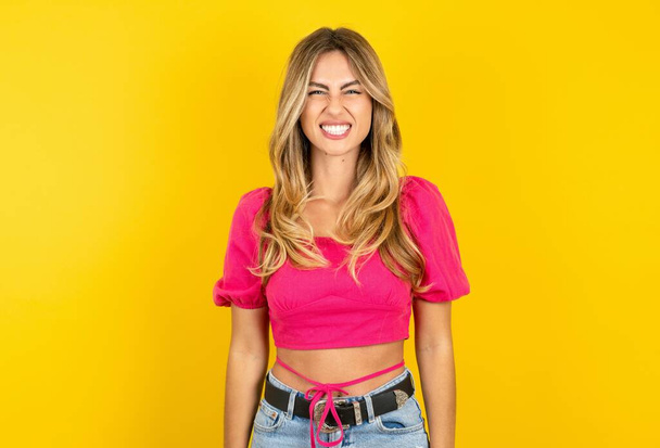 Young beautiful blonde woman wearing pink crop top over yellow  studio background keeps teeth clenched, frowns face in dissatisfaction, irritated because of much duties. - Photo, Image