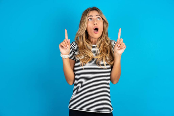 Young beautiful blonde woman wearing striped t-shirt over blue studio background amazed and surprised looking up and pointing with fingers and raised arms. - Photo, Image