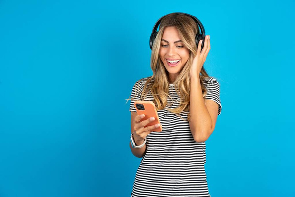 Feliz joven hermosa mujer rubia con camiseta a rayas sobre fondo de estudio azul se siente bien mientras se centra en la pantalla del teléfono inteligente. Personas, tecnología, estilo de vida - Foto, imagen
