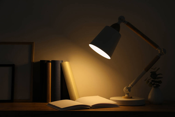 Stylish modern desk lamp, books and plant on wooden table in dark room - Foto, immagini