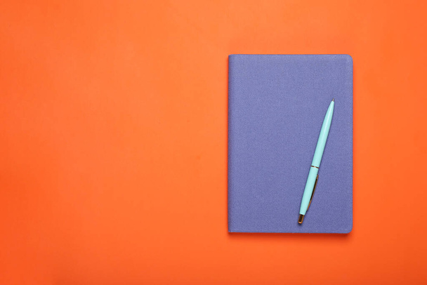 Закрытая синяя тетрадь и ручка на оранжевом фоне, вид сверху. Пространство для текста - Фото, изображение