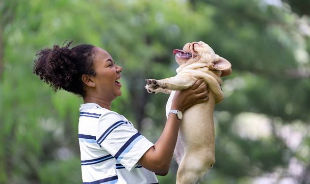 African American woman is playing with her french bulldog puppy while walking at the dog park at grass lawn after having morning exercise during summer - Photo, Image
