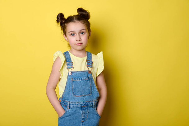 Beautiful smart clever charming little child girl, adorable school kid, pupil in yellow t-shirt and jeans overalls, holding hands in pockets, confidently looking at camera, isolated yellow background - Foto, afbeelding