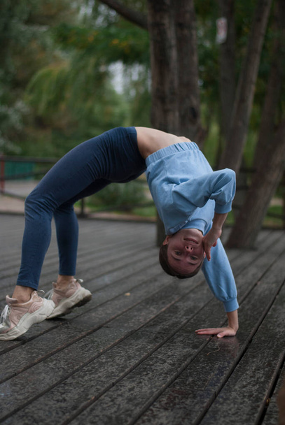 Vue de la jeune fille en costume de gymnase bleu fait des exercices acrobatiques par temps nuageux dans le parc - Photo, image