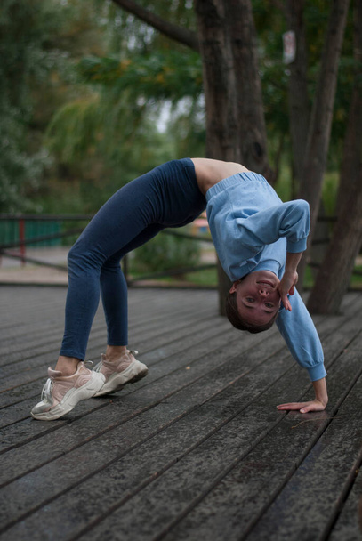 Vue sur la jeune fille en costume de gymnase bleu fait des exercices acrobatiques par temps nuageux dans le parc - Photo, image