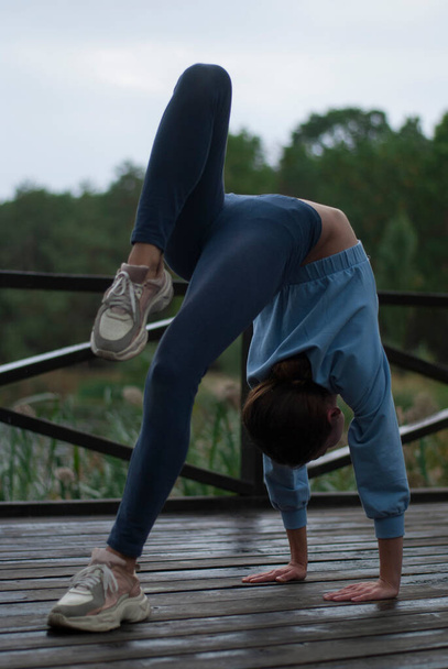 Widok dziewczyny ze sportowym ciałem w niebieskim stroju gimnastycznym robi ćwiczenia akrobatyczne w pochmurną deszczową pogodę w pobliżu lak - Zdjęcie, obraz