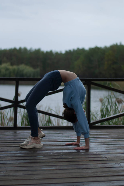 View of a girl with athletic body in blue gym suit is doing acrobatics exercises in cloudy rainy weather near lak - Photo, Image