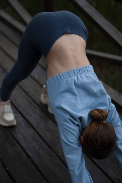 Pohled na dívku s atletickým tělem v modrém tělocvičně dělá cvičení v parku v oblačném deštivém počasí - Fotografie, Obrázek