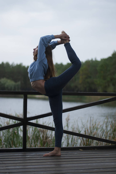 Pohled na dívku s atletickým tělem v modrém tělocvičně dělá akrobacii cvičení v oblačném deštivém počasí v blízkosti jezera - Fotografie, Obrázek