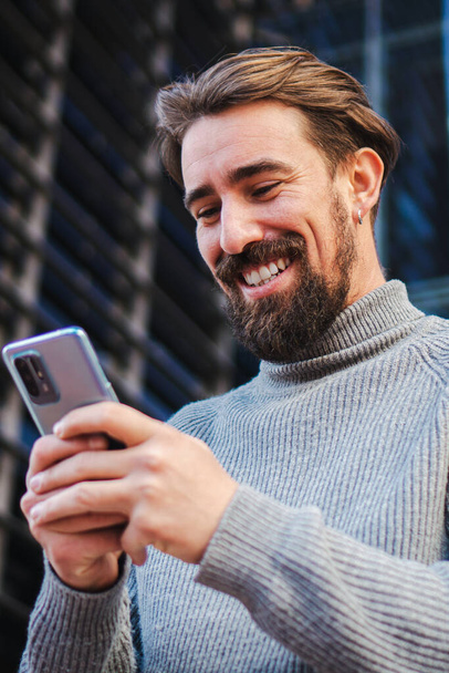 Retrato vertical de un joven hombre de negocios feliz disfrutando usando una aplicación de teléfono móvil que comparte medios en Internet. Impresionante hombre guapo mensajes de texto y riendo con el teléfono celular al aire libre. Tipo usando un teléfono móvil - Foto, imagen