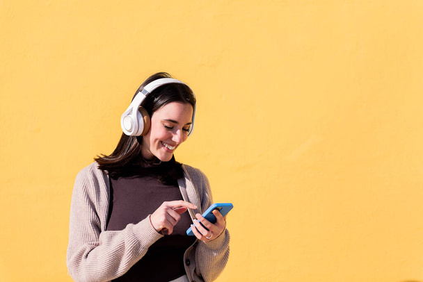 smiling young woman listening to music from phone on headphones with yellow background, concept of rhythm and positive people, copy space for text - Photo, Image