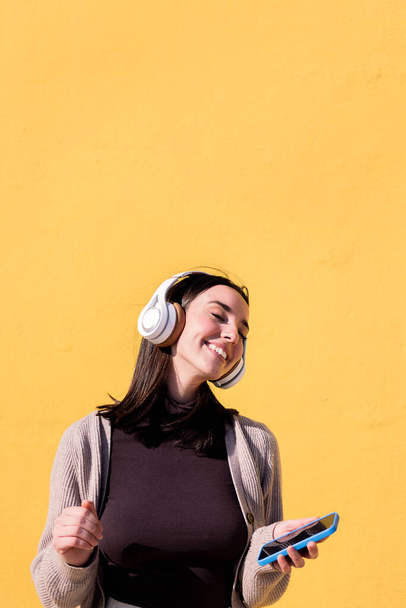 woman dancing happy listening to music from phone in headphones with a yellow wall in the background, concept of rhythm and positive people, copy space for text - Photo, Image