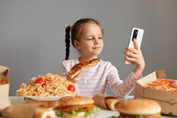 Cute little girl with braids sitting at table with junk food isolated over gray background holding slice pf pizza making selfie or video call on mobile phone. - Foto, imagen