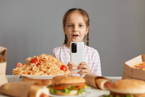Shocked astonished little girl with braids sitting at table with junk food isolated over gray background looking at camera with big eyes uaing smartp phone online food delivery. - Photo, Image