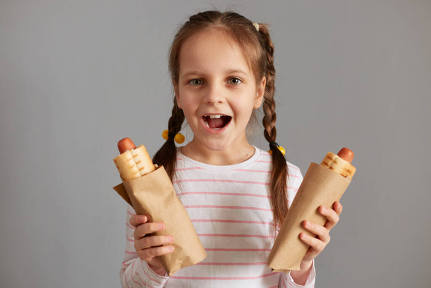Excited overjoyed little girl with pigtails with hot dogs isolated over gray background looking at camera screaming with happiness enjoyed unhealthy snack. - Fotó, kép