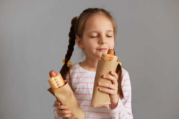 Delighted satisfied little girl with pigtails with hot dog isolated over gray background standing with closed eyes smelling aromat fresh baked fast food snack. - Fotó, kép