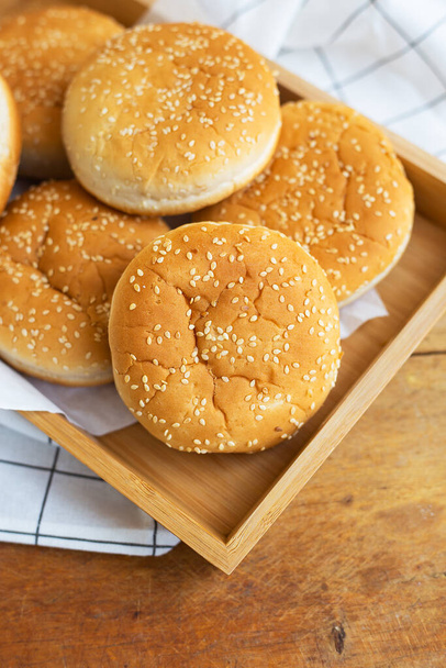 Freshly baked lush burger buns lie on a wooden tray on a table with a white napkin. Top view, close-up. Yellow flavored pastries with white sesame seeds - Fotografie, Obrázek