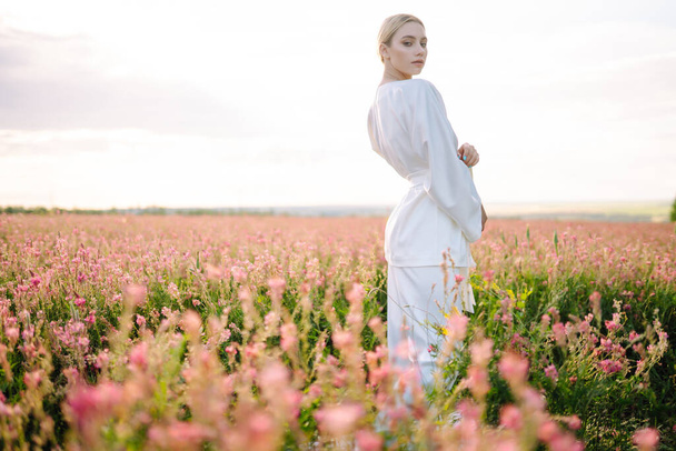 Young woman in stylish summer dress feeling free in the field with flowers in sunshine. Nature, vacation, relax and lifestyle. Summer landscape. - Фото, изображение
