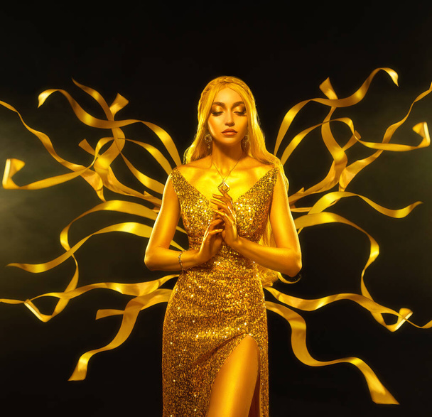 Art Fantasy woman goddess queen with metallic golden skin, long gold dress glow, ribbon flies flutters on wind motion. Night dark Black studio. Blonde fairy princess girl angel royal style clothes. - Fotó, kép