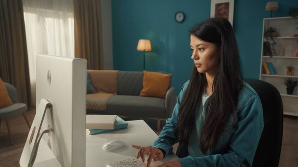 An Asian woman sits at a computer in a cozy living room. Young woman working, typing on the keyboard, writing emails, messages, chats, browsing the internet, freelancer - Φωτογραφία, εικόνα