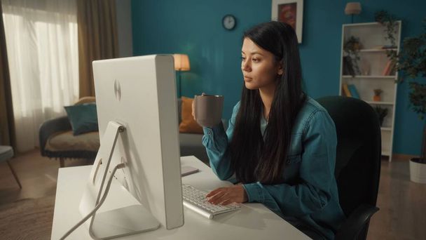 An Asian woman sits at a computer. In one hand, a woman holds a cup of coffee, the other is typing on the keyboard. Woman works, writes emails, messages, chats, surfs the internet, freelancer - Foto, afbeelding