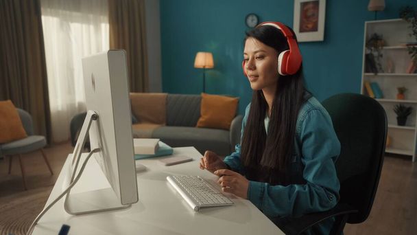 Vista lateral de una mujer asiática con auriculares inalámbricos rojos trabajando en una computadora. Una joven juega un juego en línea, trabaja, ve videos, escucha música. De cerca. - Foto, imagen